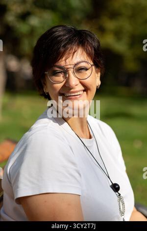 Une belle femme d'âge moyen avec des lunettes et de courts cheveux foncés est assise sur un banc Banque D'Images