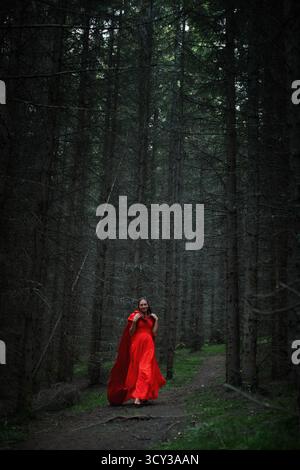 femme se promène gracieusement le long d'un chemin sinueux dans une forêt verdoyante. Elle porte une robe rouge fluide qui contraste magnifiquement avec l'obscurité, imposante Banque D'Images