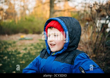 Enfant souriant en manteau d'hiver bleu et chapeau rouge à l'extérieur en automne Banque D'Images