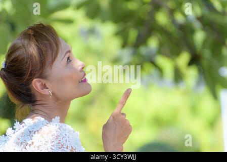 Face latérale d'une belle femme en bonne santé des gens d'Asie d'âge moyen habillent dans des vêtements de style thaïlandais, Portrait vieille dame se relaxant dehors pointant vers le doigt looki Banque D'Images