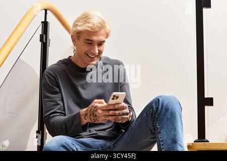 Beau jeune homme aime son temps assis dans les escaliers, engagé avec son smartphone et souriant. Banque D'Images