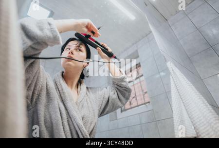 Femme utilisant l'outil de coiffure dans la réflexion de miroir de salle de bains moderne Banque D'Images