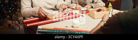 Femme caucasienne d'âge moyen et jeune femme caucasienne adulte, enveloppant des cadeaux de Noël et écrivant des cartes à table avec des rouleaux de papier d'emballage et des décorations de vacances dans un cadre festif Banque D'Images
