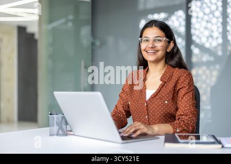 Femme d'affaires indienne souriant avec confiance. Porter des lunettes. Et taper sur un ordinateur portable tout en étant assis à un bureau propre dans un environnement de bureau moderne. Représenter le professionnalisme et la productivité Banque D'Images
