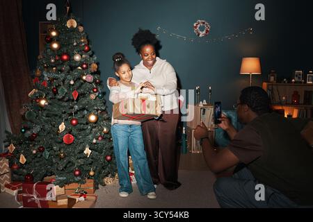 Femme noire et fille noire debout à côté de l'arbre de Noël décoré tenant des cadeaux emballés tandis que l'homme noir, agenouillé en face prendre photo avec smartphone dans le salon confortable Banque D'Images