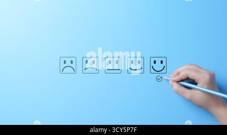 Échelle d'évaluation de la satisfaction client avec visages émoticônes. Une main marque un visage heureux sur un sondage de satisfaction client avec diverses options d'émoticônes Banque D'Images