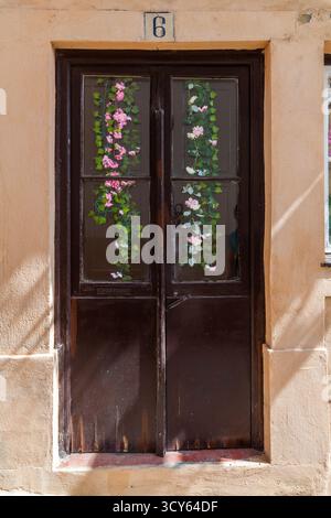 Une porte en bois sombre et vieilli avec des vitres est ornée de fleurs artificielles roses et blanches, dans un mur texturé en stuc couleur pêche Banque D'Images