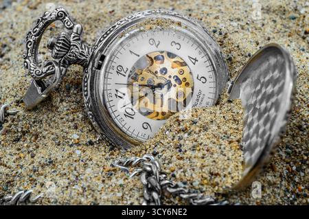 Bacau, Roumanie, 1er juillet 2015 : une montre de poche mécanique vintage aux engrenages exposés est partiellement enterrée dans le sable, évoquant des thèmes du temps et de la décomposition. Banque D'Images