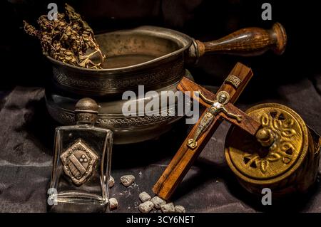 Crucifix vintage, pot gravé avec des herbes, des récipients ornés et des pierres dispersées disposées sur un tissu sombre. Banque D'Images