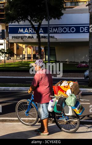 Marilia, SP, Brésil, 1er mai 2025. Homme âgé conduisant un vélo chargé devant le Conseil municipal et l'Hôtel de ville sous la phrase : symbole de l'amour et FR Banque D'Images