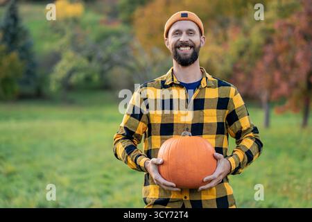 Portrait d'un jeune homme à la barbe dans un chapeau orange dans un paysage d'automne. tenant une grande citrouille dans ses mains. Banque D'Images