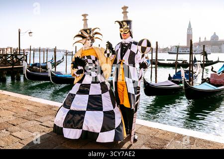 Costumes de carnaval de Venise sur le front de mer avec gondoles. Deux participants masqués en costumes noir, blanc et or posent près du Grand canal. Banque D'Images