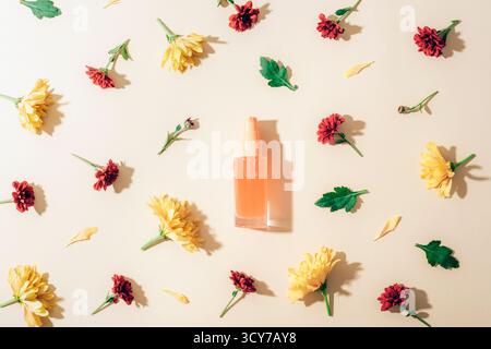 Bouteille cosmétique en verre avec des chrysanthèmes jaunes et rouges sur fond beige avec des ombres. Flat lay, concept de beauté naturelle. Vue de dessus, maquette. Banque D'Images