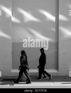 Trottoir monochromatique ; silhouettes contrastées avec un mur texturé avec des puits de lumière du soleil ; sentiment d'humeur changeante de la vie urbaine anonyme. Banque D'Images