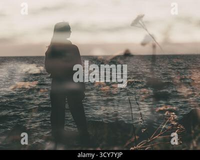 Femme silhouettée par la mer, contemplant l'horizon au coucher du soleil. Une fille rêveuse pensive se tient sur le rivage, regardant l'océan sous un au moody Banque D'Images