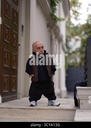 Homme avec le nanisme debout sur les marches à l'extérieur de la maison dans le sweat à capuche et gilet, faire pouting ou expression insatisfaite. Personne unique près de la porte en bois. Banque D'Images