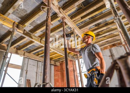 Un travailleur de la construction dans un casque de sécurité ajuste les supports d'échafaudage dans un bâtiment en construction pendant la journée. Banque D'Images