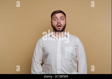 Un homme avec une barbe se tient sur un fond Uni portant une chemise blanche. Ses yeux sont larges et la bouche ouverte, exprimant la surprise ou l'incrédulité. Banque D'Images