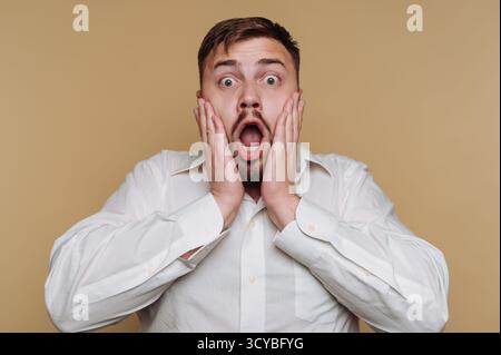 Un homme portant une chemise blanche affiche une expression choquée avec les yeux et la bouche grands ouverts. Il a les mains pressées contre son visage, capturant un sens Banque D'Images