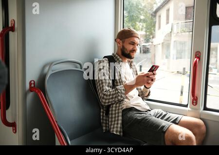 Jeune homme assis dans un bus de la ville, regardant son smartphone en voyage. Concept de communication, de technologie et de mobilité urbaine. Banque D'Images