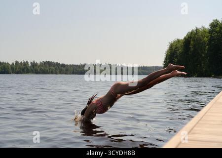 Une femme en maillot de bain se tient sur une jetée au bord du lac avec les bras tendus, embrassant le soleil et l'air frais. Moment paisible de liberté, de nature et de connexion avec le corps. Banque D'Images