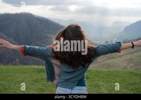 Vue arrière de la jeune femme en short et chemise debout au bord de la montagne, les bras tendus dans des conditions venteuses. Le temps nuageux rehausse les paysages spectaculaires. Banque D'Images