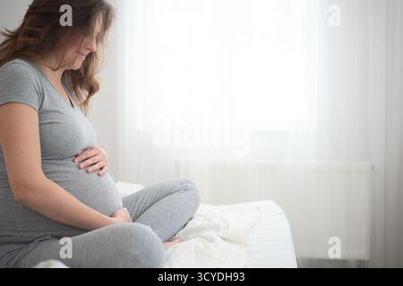 Femme enceinte brillante assise confortablement sur le lit tout en touchant tendrement son ventre. Scène réconfortante de bonheur maternel et d'attente joyeuse. Banque D'Images