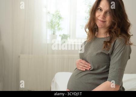 Femme enceinte rayonnante assise sur le lit, touchant amoureusement son ventre dans un cadre domestique confortable. Belle anticipation de la maternité et nouvelle vie. Banque D'Images