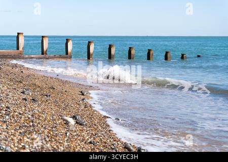De douces vagues se jettent sur une plage de galets avec une ligne de groynes en bois altérés s'étendant dans la mer bleue calme sous un ciel clair. Banque D'Images