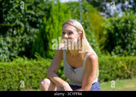 Une femme blonde s'accroupit à l'extérieur dans un jardin, souriant brillamment à la lumière du soleil tout en profitant de la journée d'été. Banque D'Images