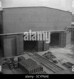 Cette photo d'archives en noir et blanc des années 1970 capture une vue en grand angle d'un grand atelier de briques à l'usine d'armature-isolant Slavyansk (AIZ) à Sloviansk, URSS. Deux portes massives en bois du bâtiment sont grandes ouvertes. Des voies ferrées sont visibles menant directement à l'entrée gauche, un élément crucial de la logistique de l'usine pour le transport des matières premières ou des produits finis. Au premier plan, la cour est utilisée comme zone de stockage temporaire, avec des milliers d'isolants finis soigneusement empilés en grandes piles, mettant en valeur la production de l'usine. Cette image illustre l'infrastructure industrielle Banque D'Images
