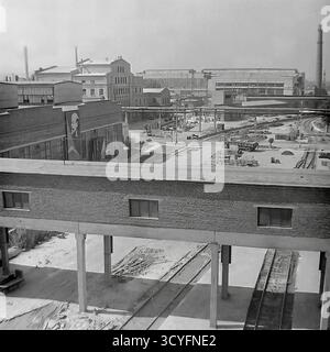 Cette photo d'archives en noir et blanc des années 1970 donne une vue en grand angle de l'armature-isolant Slavyansk (AIZ) à Sloviansk, URSS. La photo donne sur une grande passerelle couverte surélevée (pont piétonnier) qui traverse plusieurs voies ferrées en contrebas. En arrière-plan, la vaste cour d'usine est visible, animée de camions, de travailleurs et de zones de stockage de produits. Un grand portrait de Vladimir Lénine est bien visible sur le mur d'un atelier. Cette image montre l'infrastructure complexe d'un site industriel soviétique majeur Banque D'Images