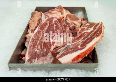 Des coupes épaisses et crues de viande rouge non désossée sont exposées dans un plateau métallique reposant sur un lit de glace pilée dans une boucherie ou un comptoir de supermarché. Banque D'Images