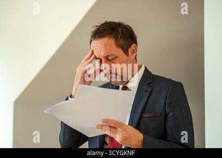 Homme d'affaires inquiet en costume lisant des documents financiers, souligné par le rapport négatif, examinant la paperasse et souffrant de stress au travail. Sélectif Banque D'Images
