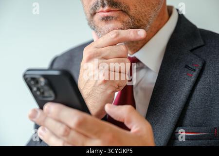 Homme d'affaires contemplatif tenant un smartphone et lisant un message texte avec la main sur le menton, moment de bureau réfléchi. Mise au point sélective. Banque D'Images