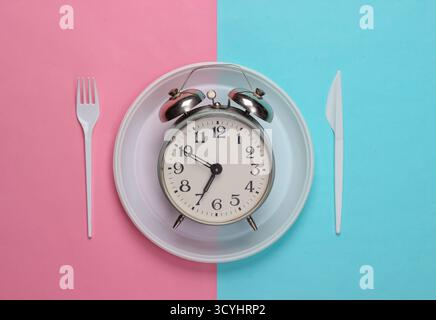 Heure du pique-nique. Réveil dans une assiette avec une fourchette jetable et un couteau sur fond pastel bleu rose. Vue de dessus Banque D'Images