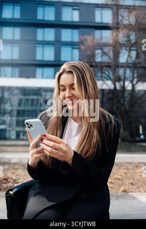 Une jeune femme est assise sur un banc, s'engageant joyeusement avec son smartphone dans un quartier moderne de la ville. Les grands bâtiments offrent une toile de fond élégante sous un SK bleu clair Banque D'Images