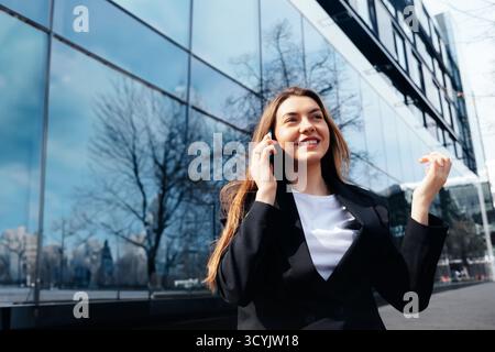 Une jeune femme vêtue d'une tenue professionnelle parle sur son téléphone tout en se tenant debout à l'extérieur d'un élégant immeuble de bureaux, souriant et faisant des gestes pendant qu'elle m'engage Banque D'Images