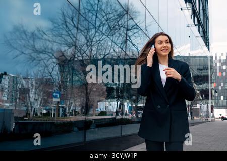 Une jeune femme se promène le long d'une passerelle pavée à côté d'un bâtiment élégant, s'engageant dans une conversation téléphonique tout en profitant du temps clair au milieu des s urbains Banque D'Images