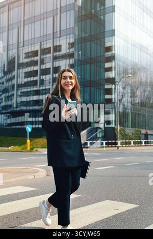 Une jeune femme confiante marche dans une rue urbaine, vêtue d'un costume noir, souriant en utilisant son smartphone devant un immeuble de bureaux moderne. Banque D'Images