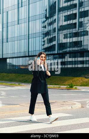 Une femme vêtue d'une tenue d'affaires marche en toute confiance à travers un passage pour piétons tout en parlant au téléphone. Les bâtiments modernes reflètent son look élégant sur un su Banque D'Images