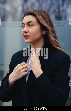 Une jeune femme se tient confiante dans un environnement urbain, ajustant sa veste tout en affichant une expression sereine sur un fond doux de ville Ar Banque D'Images