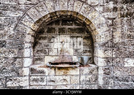 Ancienne fontaine en pierre construite dans un mur voûté à Prague, en République tchèque, avec une coupe en métal reposant à côté du bassin usé. Banque D'Images