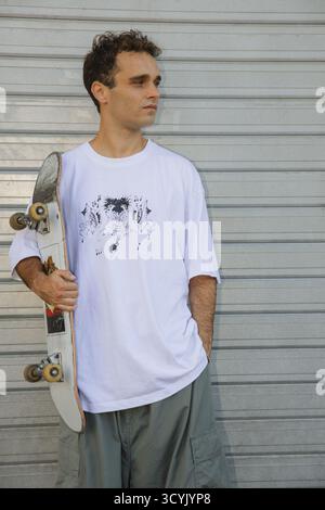 Un jeune patineur élégant tient son skateboard en toute confiance contre un fond d'obturateur métallique. Banque D'Images