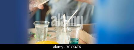 Mains fabriquant de l'art en résine coloré avec des bâtons de mélange et des gobelets en plastique transparent dans un cadre de studio. Banque D'Images
