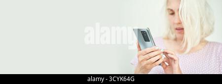 Femme adulte caucasienne blonde à l'aide d'un smartphone dans une chemise à pois rose clair sur fond Uni. Banque D'Images