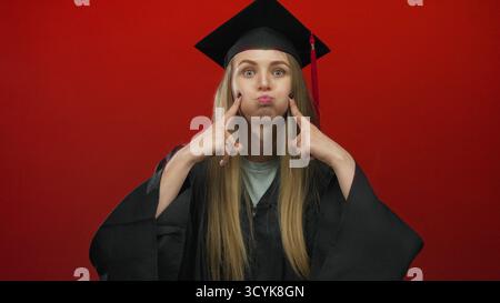 Une jeune femme vêtue d'une tenue de remise des diplômes se dresse contre un mur rouge vif, exprimant une émotion ludique, mettant en valeur une vision légère de l'academ traditionnel Banque D'Images
