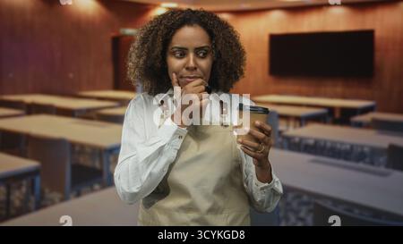 Femme dans le tablier tient une tasse de café et touche le menton dans la salle de classe de construction tout en considérant une note de recette ; curiosité réfléchie. Banque D'Images