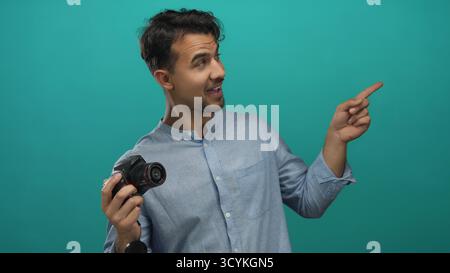 Jeune homme hispanique tenant une caméra pointant quelque chose sur fond vert isolé suggérant une idée ou une direction Banque D'Images