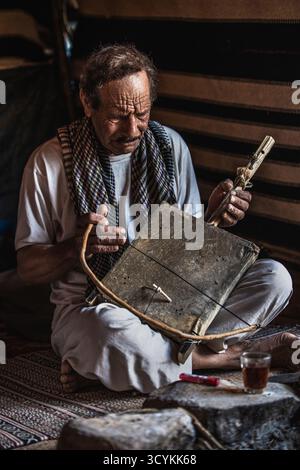 Homme bédouin portant une écharpe traditionnelle, jouant de la rababa, un instrument à cordes traditionnel, dans une tente dans le désert de Wadi Rum, Jordanie. 2024 Banque D'Images